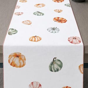 May include: A white table runner featuring a repeating pattern of watercolour-style pumpkins in various shades of orange, sage green, and muted pink. The rectangular runner has a thin, solid-coloured border along its long edges and a smooth finish.