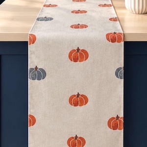 May include: A rectangular table runner made of light beige fabric featuring a repeating pattern of printed pumpkins. The pumpkins are coloured in shades of burnt orange and muted slate blue, arranged in a grid-like layout across the entire surface.