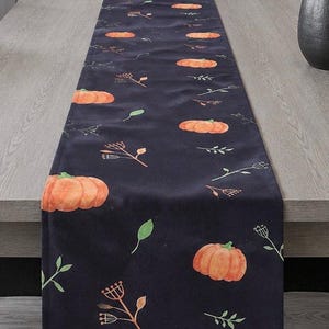 May include: A long, rectangular table runner in a deep black fabric featuring a repeating pattern of orange pumpkins, small green leaves, and delicate brown botanical sprigs. The runner is laid flat across a light-coloured wooden dining table surface.