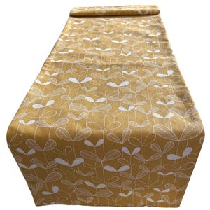 May include: Mustard yellow table runner featuring a repeating white leaf pattern. The runner is made of a textured fabric and is folded at the top. The leaf design is a simple, stylized botanical print.