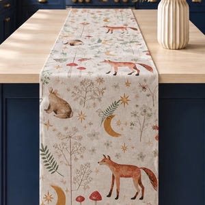 May include: A rectangular table runner featuring a repeating pattern of orange foxes, brown rabbits, red-capped mushrooms, golden crescent moons, and stars on a light beige fabric background. The design includes various botanical illustrations.