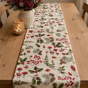May include: A cream-coloured table runner featuring a festive pattern of red mushrooms, berries, and green foliage. The runner is on a wooden table, creating a seasonal display. A vase of red berries and a candle holder are also on the table.