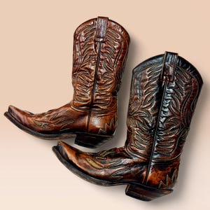 Puede incluir: Un par de botas de vaquero de cuero marrón con intrincados diseños florales y de hojas. Las botas tienen puntas puntiagudas y tacones apilados, mostrando un estilo occidental clásico. El cuero tiene un aspecto desgastado.