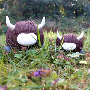 May include: Two brown Highland cow figurines with white horns and white muzzles. The larger figurine is on the left and the smaller one is on the right. Both figures have textured brown hair and are positioned side by side in a grassy outdoor area.