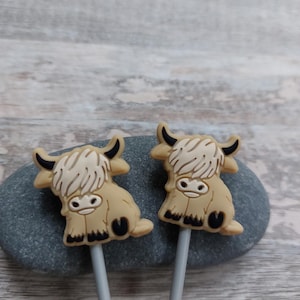 May include: Two Highland cow-shaped toppers with tan bodies, black horns, and white fur accents. The toppers are attached to grey sticks. The cows are resting on a grey stone against a light wood-grain background.