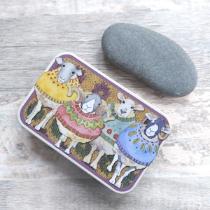 May include: A rectangular tin with an illustration of four sheep in patterned sweaters. The tin has a purple border and is next to a smooth, grey stone. The tin is on a light wood surface. The sheep are wearing sweaters in various colours.