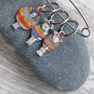 May include: Three decorative sheep stitch markers with silver rings and a safety pin. The sheep are adorned with colourful patterned coats. The markers are displayed on a grey stone, with a wooden background.