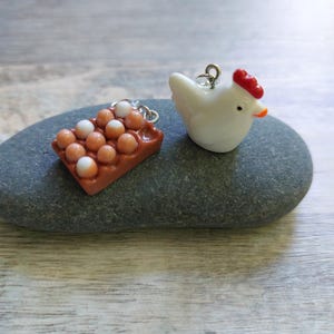 May include: Two miniature charms: a white chicken with a red comb and orange beak, and a brown egg carton with white and brown eggs. Both charms have silver-coloured loops for hanging. The charms are on a grey stone.