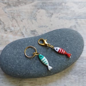 May include: Two fish-shaped keyrings and a charm. One keyring has a gold ring and a teal and white fish charm. The other has a gold clasp and a red and white striped fish charm. The charms are on a grey stone.