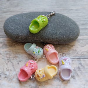 May include: A selection of miniature clog charms in green, pink, yellow, and blue. Each charm has a silver loop for attaching to a keychain or bracelet. The charms are displayed on a grey stone. Metric measurements.