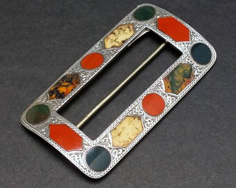 May include: A rectangular silver buckle with a central bar. The buckle is adorned with a variety of coloured stones, including red, green, and cream-coloured stones. The edges of the buckle have a detailed, patterned design.