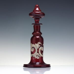 Puede incluir: Una botella de vidrio rojo rubí con un tapón decorativo. La botella presenta un diseño plateado, ornamentado y arremolinado alrededor de la base. La botella mide aproximadamente 25 cm de altura.