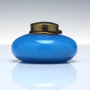 Puede incluir: Pequeña botella de vidrio azul claro de forma ovalada, con tapa color latón. La botella tiene un acabado liso y brillante y un aspecto ligeramente moteado. La tapa es circular y encaja perfectamente. Probablemente un frasco de perfume.