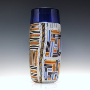 May include: A tall, cylindrical glass vase with a blue top and a colorful geometric pattern. The pattern is made up of stripes, squares, and other shapes in shades of orange, yellow, white, black, and blue.