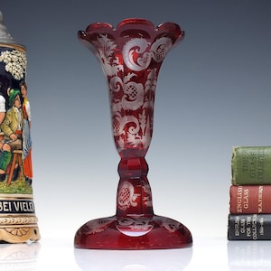 Puede incluir: Un jarrón de cristal rojo rubí con diseños florales y de volutas grabados. El jarrón tiene una parte superior acampanada y una base ancha y circular. Una jarra de cerveza de cerámica y una pila de libros están en el fondo.