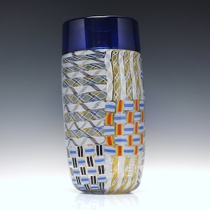May include: A tall, cylindrical glass vase with a blue rim and a colorful geometric pattern. The pattern features white, blue, yellow, orange, and black stripes and squares.