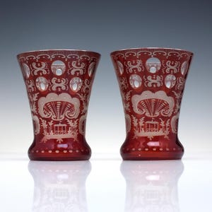 May include: A pair of ruby red glass vases with detailed etched patterns. The vases have a flared shape, with intricate designs of floral and geometric elements. The etched patterns are in a lighter tone, creating a contrast against the red glass.