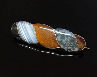 May include: A silver brooch with a twisted design featuring four different types of agate stones. The stones are a mix of white, brown, green, and red.