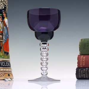May include: A deep purple glass wine goblet with a clear glass stem featuring a beaded design. The goblet is sitting on a white surface.
