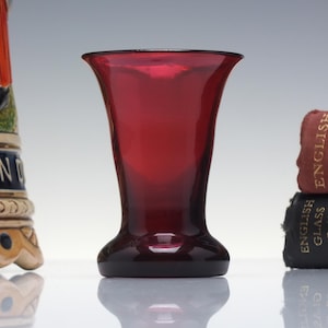 May include: A deep ruby red glass vase with a flared rim and a smooth, tapered body that widens towards the top. The vase features a thick, solid base and a translucent finish that allows light to pass through the coloured glass material.