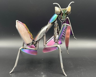 May include: A metallic praying mantis sculpture with iridescent purple, pink, and green accents. The sculpture is made of recycled metal and has a unique, abstract design.