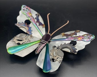 May include: A metal butterfly sculpture with iridescent wings made from repurposed gears and other metal pieces. The wings are a mix of silver, green, and purple.