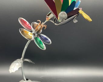 May include: A colorful metal hummingbird sculpture with a multi-colored flower, perched on a metal stand. The hummingbird is made from recycled materials and has a unique, whimsical design.