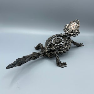 May include: A metal sculpture of a lizard made from bike chains and gears. The lizard is in a standing position with its tail extended behind it.