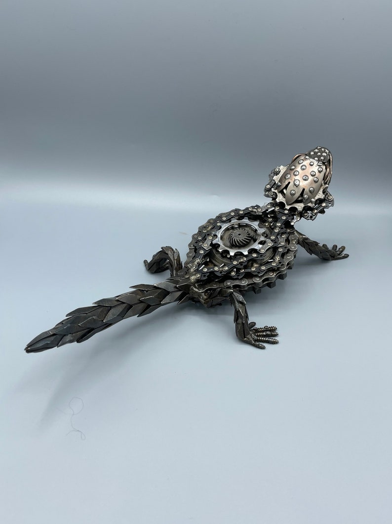 May include: A metal sculpture of a lizard made from bike chains and gears. The lizard is in a standing position with its tail extended behind it.