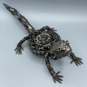 May include: A metal sculpture of a lizard made from recycled bike chains and other metal parts. The lizard is posed in a walking position with its head turned to the side. The sculpture is painted in a silver color.