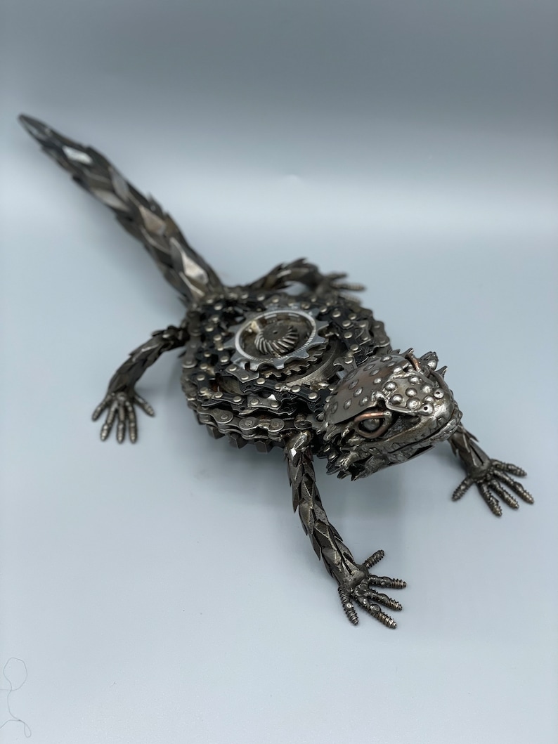 May include: A metal sculpture of a lizard made from recycled bike chains and other metal parts. The lizard is posed in a walking position with its head turned to the side. The sculpture is painted in a silver color.