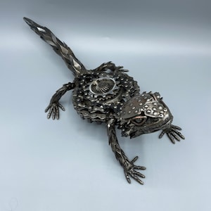 May include: A metal sculpture of a lizard made from recycled metal parts, including chains and gears. The lizard is posed in a walking position with its head turned to the side. The sculpture is painted in a metallic silver color.
