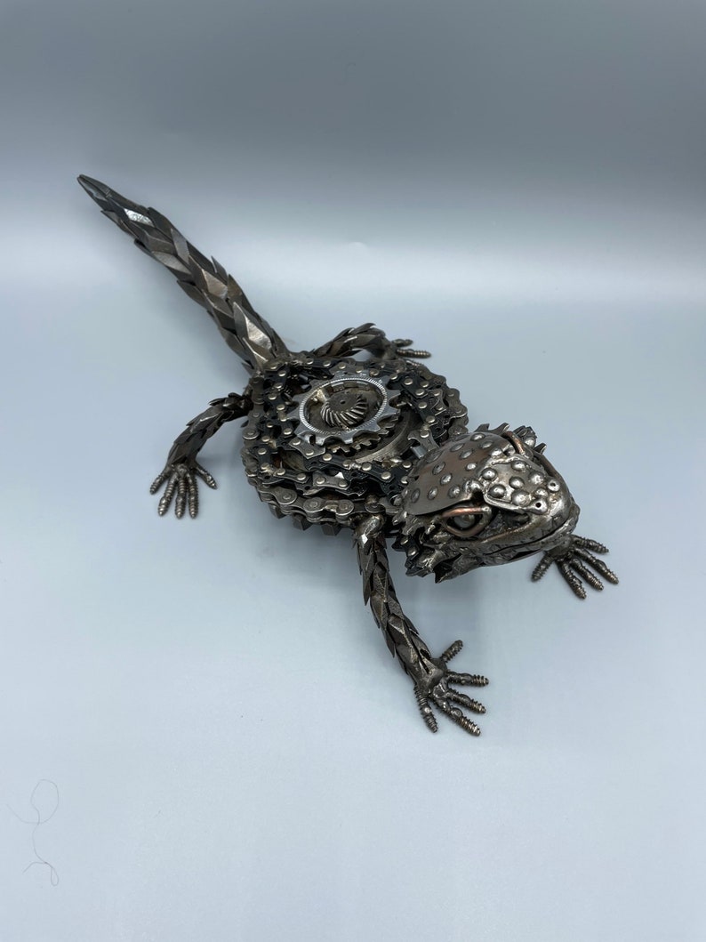 May include: A metal sculpture of a lizard made from recycled metal parts, including chains and gears. The lizard is posed in a walking position with its head turned to the side. The sculpture is painted in a metallic silver color.