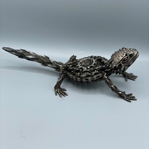May include: A metal sculpture of a lizard made from recycled bike parts. The lizard is posed in a walking position with its tail extended behind it. The sculpture is made of silver and black metal.