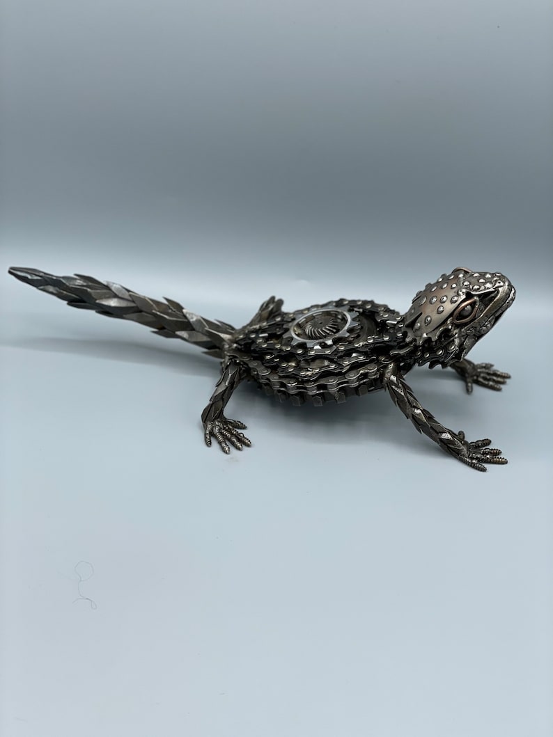 May include: A metal sculpture of a lizard made from recycled bike parts. The lizard is posed in a walking position with its tail extended behind it. The sculpture is made of silver and black metal.