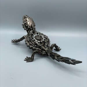 May include: A metal sculpture of a lizard made from bike chains and gears. The lizard is posed in a walking position with its tail extended behind it.