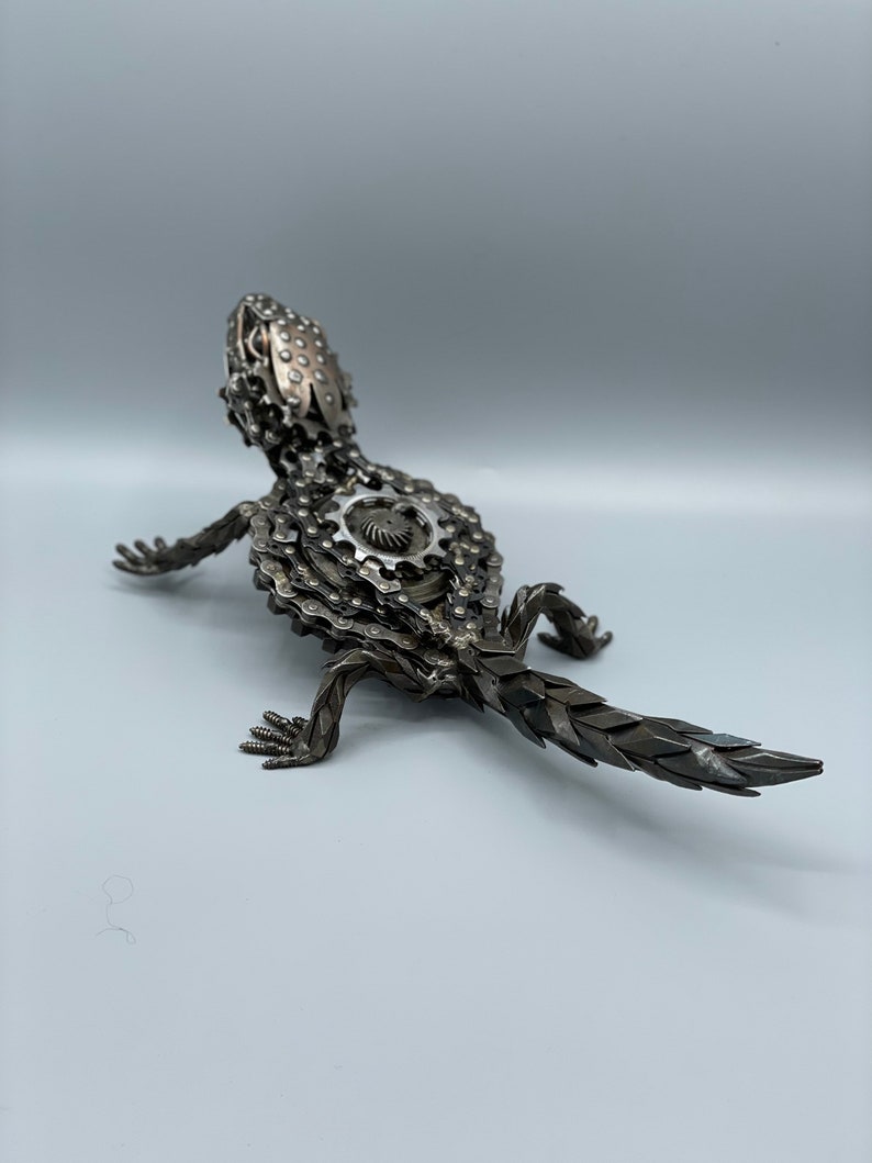 May include: A metal sculpture of a lizard made from bike chains and gears. The lizard is posed in a walking position with its tail extended behind it.