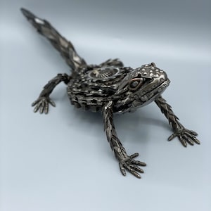 May include: A metal sculpture of a lizard made from recycled bike parts. The lizard is posed in a walking position with its tail extended behind it. The sculpture is painted in a metallic silver color.