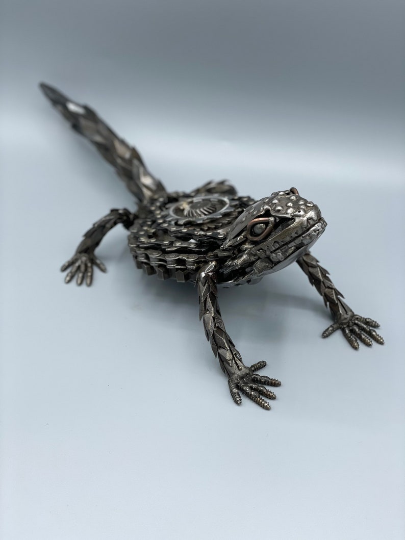 May include: A metal sculpture of a lizard made from recycled bike parts. The lizard is posed in a walking position with its tail extended behind it. The sculpture is painted in a metallic silver color.