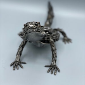 May include: A metal sculpture of a horned lizard, made from recycled materials. The lizard is posed in a crawling position with its head raised and its eyes looking directly at the viewer.