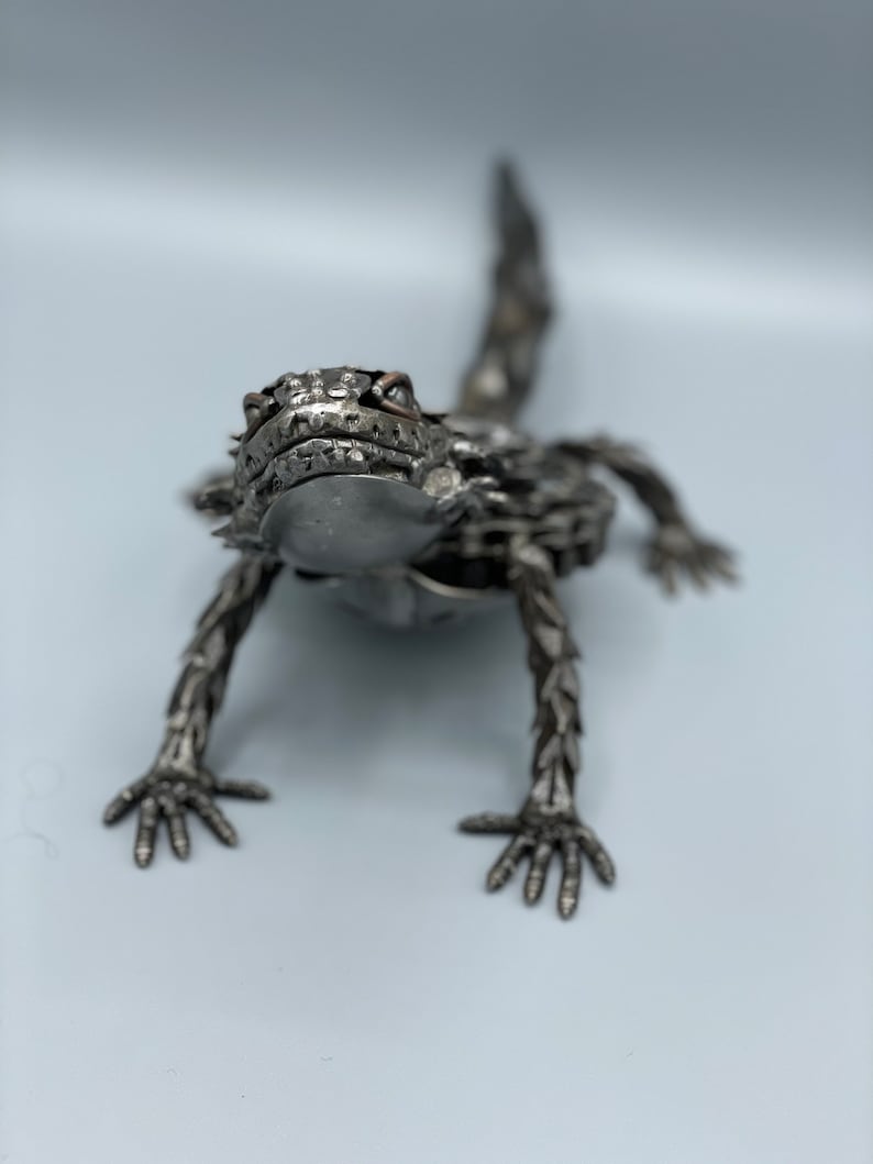 May include: A metal sculpture of a horned lizard, made from recycled materials. The lizard is posed in a crawling position with its head raised and its eyes looking directly at the viewer.