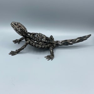 May include: A metal sculpture of a lizard made from recycled metal parts, such as chains and gears. The sculpture is painted in a metallic silver color and has a realistic appearance.