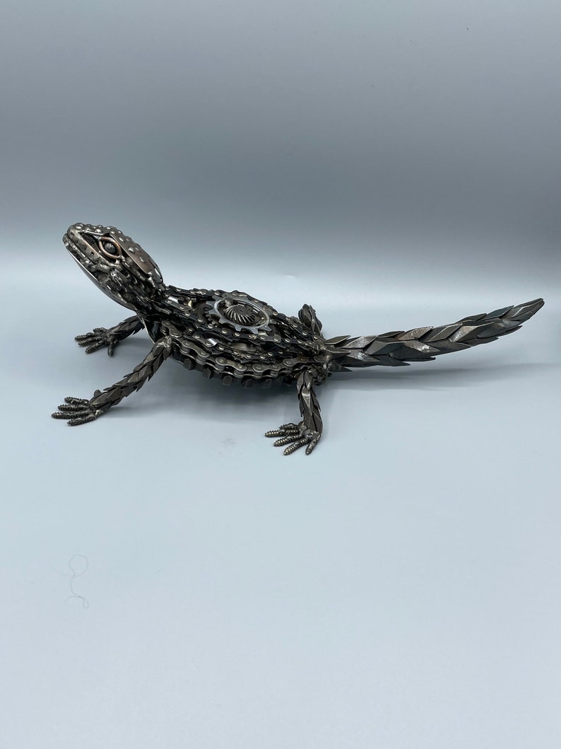 May include: A metal sculpture of a lizard made from recycled metal parts, such as chains and gears. The sculpture is painted in a metallic silver color and has a realistic appearance.
