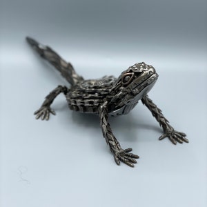 May include: A metal sculpture of a lizard, made from recycled materials. The lizard is posed in a walking position with its head raised and its tail extended behind it. The sculpture is painted in a silver color.