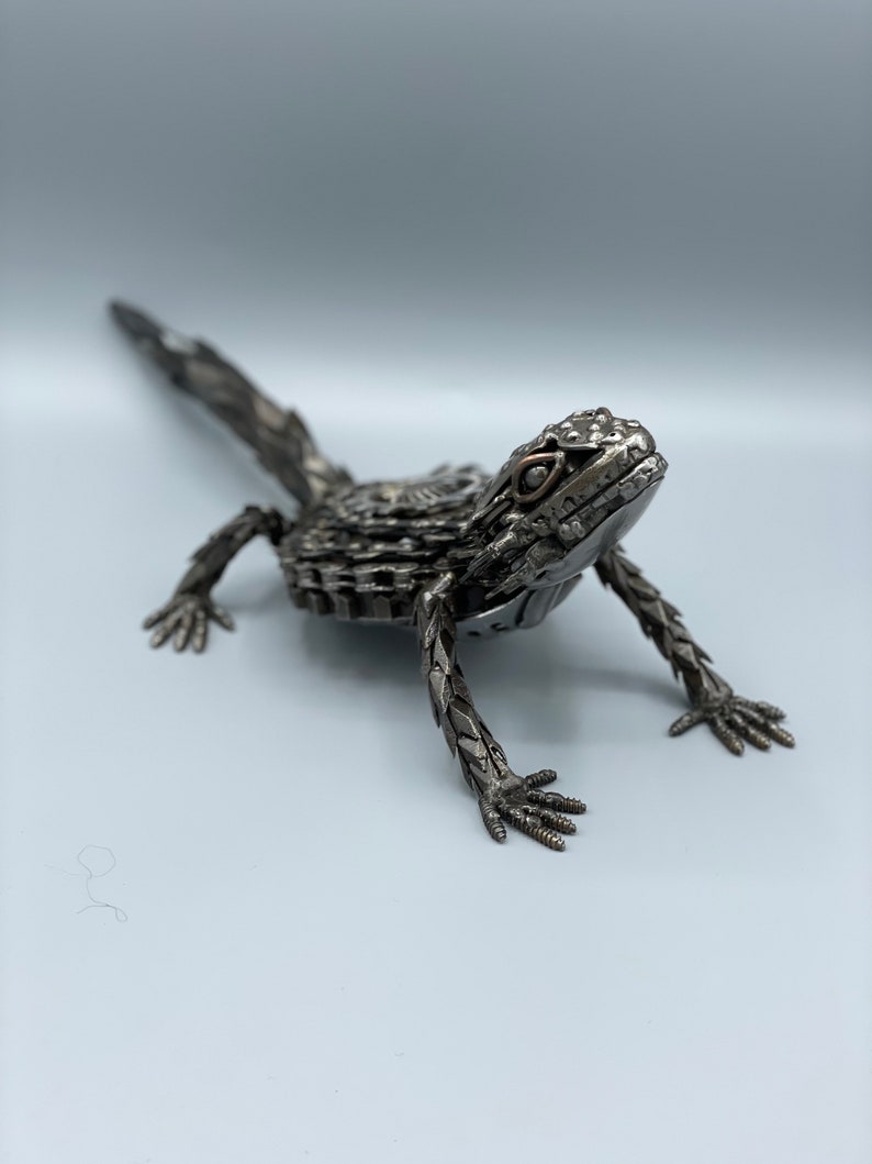 May include: A metal sculpture of a lizard, made from recycled materials. The lizard is posed in a walking position with its head raised and its tail extended behind it. The sculpture is painted in a silver color.
