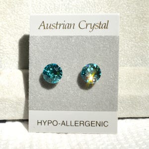 May include: Two round, teal-colored Austrian crystal stud earrings. The earrings are displayed on a gray card with the text "Austrian Crystal" and "HYPO-ALLERGENIC". The crystals have a faceted design.