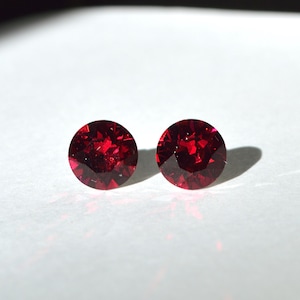 May include: Two round, faceted, deep red gemstones, possibly rubies or garnets, are displayed against a white background. The gems are circular, with multiple facets catching the light, creating a sparkling effect. The image is a close-up, highlighting the color and clarity of the stones.
