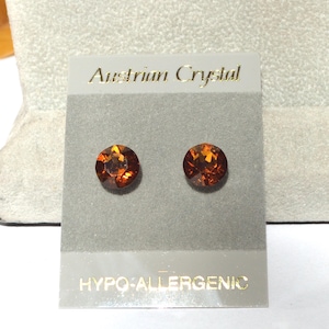 May include: A pair of round, orange-brown Austrian Crystal stud earrings. The earrings are displayed on a gray card with the words "Austrian Crystal" and "HYPO-ALLERGENIC" printed in gold lettering.