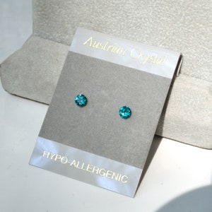 May include: A pair of small, round, teal blue Austrian crystal earrings on a white card that reads "Austrian Crystal" and "Hypo-allergenic".