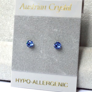 May include: A pair of small, blue, round stud earrings with a silver backing. The earrings are on a white card with the text "Austrian Crystal" and "Hypo-allergenic" printed in gold.